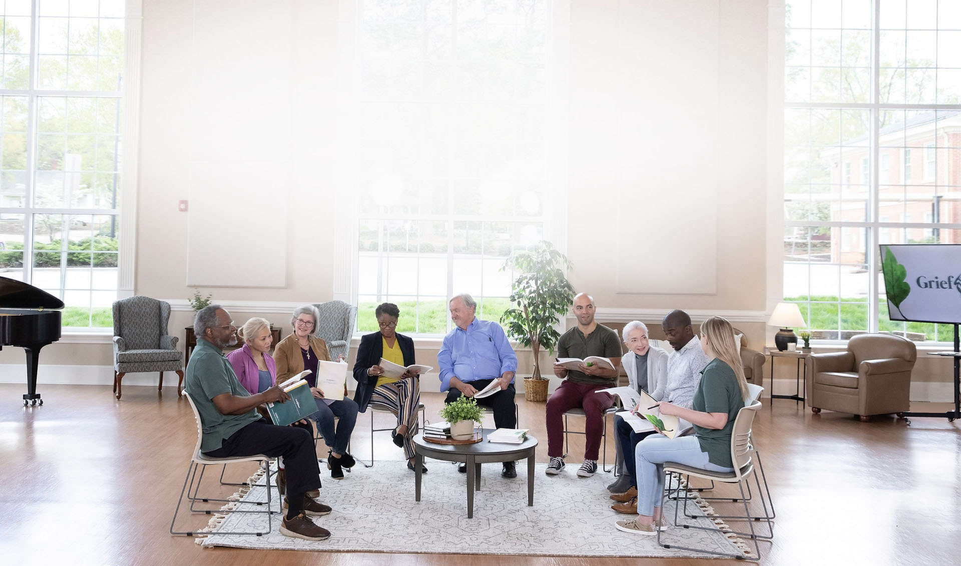 A diverse group of people sit together at a GriefShare support group. They are holding workbooks and warmly interacting.